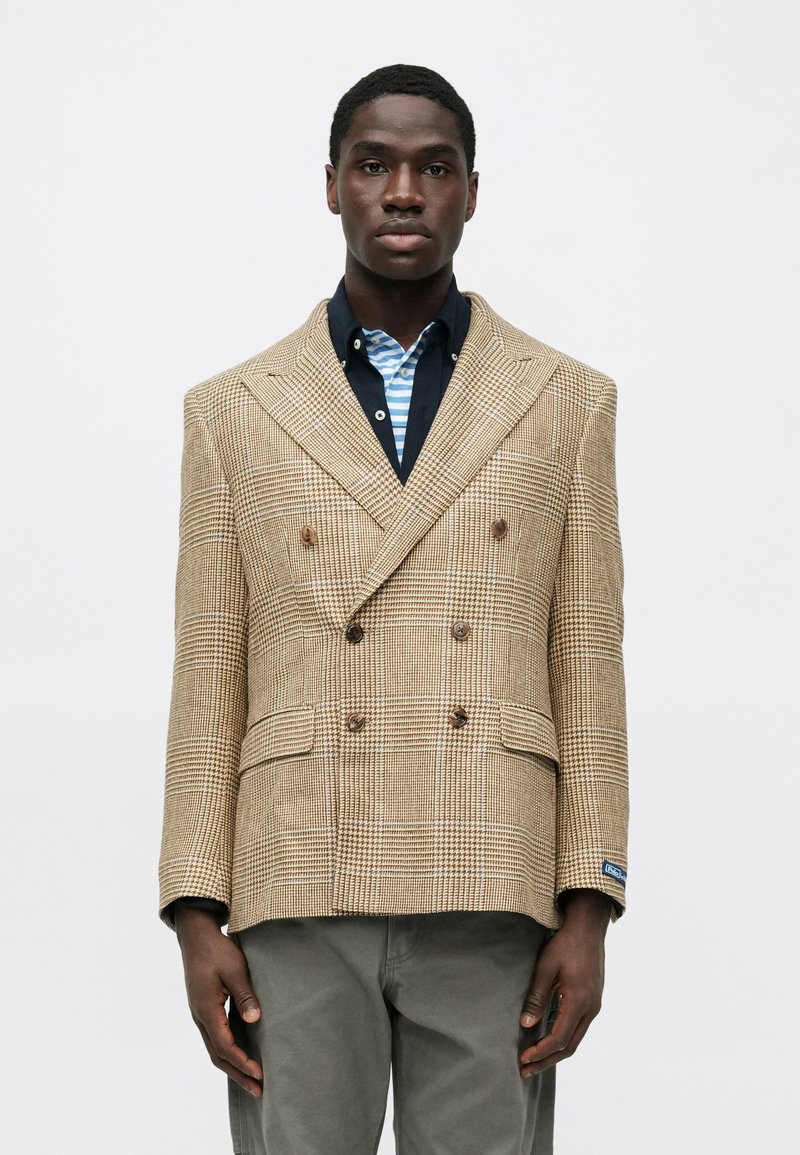 Bež dvoredni sako na kockice, s smeđim gumbima, bočnim džepovima i šiljatim reverima, nošen preko prugaste košulje.