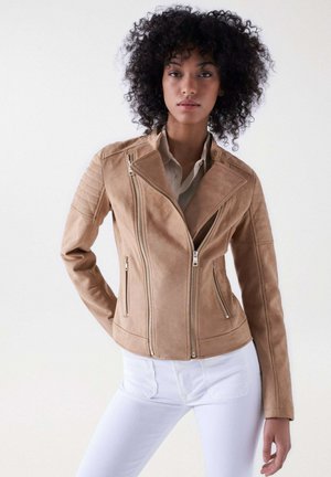 Veste de moto en fausse suède beige avec fermeture éclair diagonale, poches latérales et accents matelassés aux épaules. Portée sur une chemise crème et un jean blanc.