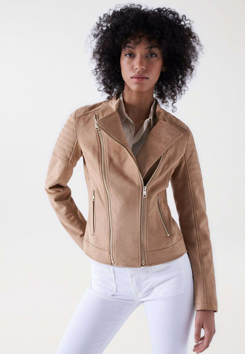 Veste de moto en fausse suède beige avec fermeture éclair diagonale, poches latérales et accents matelassés aux épaules. Portée sur une chemise crème et un jean blanc.