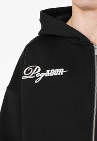 Crna zip-up dukserica napravljena od mekanog materijala. Sadrži izvezeni natpis "Pegarbor" u bijeloj boji na lijevoj strani prsnog područja. Dizajn s kapuljačom i opušteni kroj.