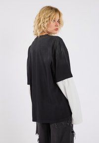 Übergroßes schwarzes T-Shirt mit einer weißen Langarmunterlage, das eine weiche Textur und einen lässigen Schnitt aufweist. Ausgewaschene schwarze Jeans runden den Look ab.