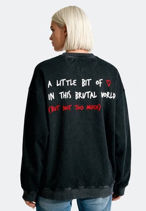 Donna con capelli biondi corti che indossa una felpa nera con la scritta "A little bit of love in this brutal world (but not too much)" in bianco e rosso.