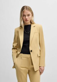 Yellow blazer and matching trousers in a smooth fabric. Features a single button closure, notch lapels, and side pockets. Black top underneath.