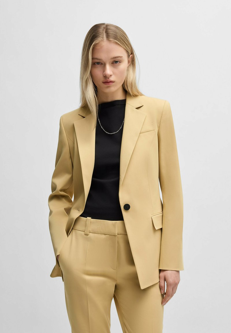Yellow blazer and matching trousers in a smooth fabric. Features a single button closure, notch lapels, and side pockets. Black top underneath.