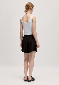 Witte tanktop met een ronde rug, gecombineerd met zwarte, elastische shorts. Model draagt zwarte instapsandalen en kijkt van de camera weg.