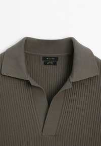 Ribbet, olivengrøn polo-shirt med klassisk krave og vævet tekstur. Har et mærke med brand og plejeinformation.