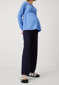 Blå texturerad graviditetströja med empire midja, i kombination med marinblå vida byxor och svarta sandaler med vita remmar och spänne.