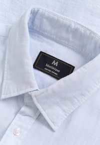 Hellblaues Hemd mit Kragen aus strukturiertem Stoff. Es verfügt über ein schwarzes Etikett mit goldener Stickerei, das den Markennamen "Matinique" zeigt.