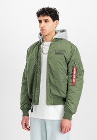 Giacca bomber verde oliva con polsini e orlo a costine, dotata di tasca con zip e un'etichetta rossa. Sovrapposta a una felpa grigia con cappuccio all'interno.