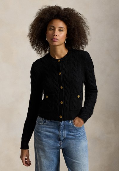 Sort kabelstrikket cardigan med guldknapper, der har to forlommer foran. Stoffet fremstår tekstureret og kombineres med lyseblå jeans.