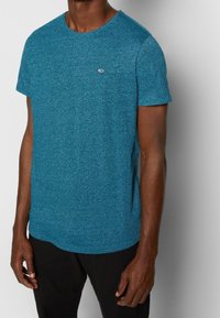 Tričko s krátkým rukávem vyrobené z texturované tyrkysové tkaniny, s kulatým výstřihem a malým logem na hrudi. Rovný lem.
