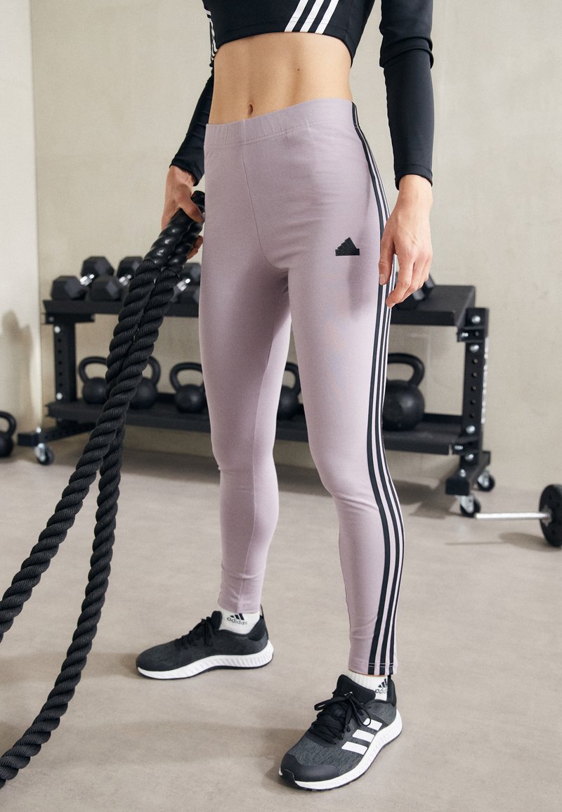 Leggings sportivi di un violetto chiaro con strisce nere laterali e logo Adidas. Il tessuto è liscio con un design aderente, abbinati a sneakers nere.