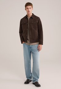 Rjava suedina jakna z dvema sprednjima žepoma, nošena čez sivo majico. V kombinaciji z svetlim modrim širokim hlačami in črnimi gležnarji.