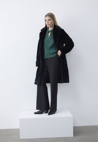 Vrouw staand op een wit platform, gekleed in een zwarte bontjas, groene blouse, zwarte brede broekspijpen en zwarte laarzen met hakken, tegen een witte muur.