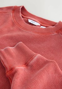 Sweat-shirt rouge texturé avec un col et des poignets côtelés. Étiquette de la marque visible à l'intérieur du col.