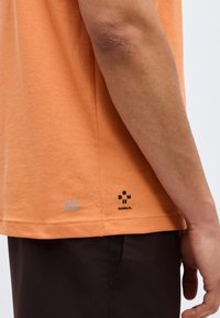 Πλάγια όψη ενός ατόμου που φοράει πορτοκαλί μπλουζάκι με τα λογότυπα "ULTRA DRY" και "DANHILM." στο κάτω μέρος και μαύρο παντελόνι.