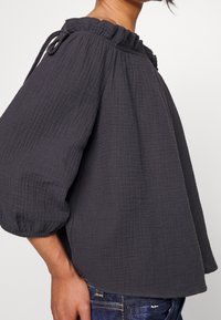 Grå texturerad blus med volangkrage och dragsko; har lösa, luftiga ärmar och en avslappnad passform.