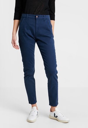 Personne portant un pantalon slim bleu marine, un haut noir à manches longues et des baskets blanches, debout devant un fond uni.