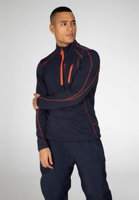 Fleece blu navy con zip, dettagli rossi, colletto alto, maniche lunghe e tasca sul petto. Texture liscia, vestibilità atletica e cuciture a contrasto.