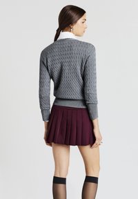 Donna in piedi di lato che indossa un maglione grigio a trecce, una camicia bianca col colletto, una gonna plissettata bordeaux e calze nere alti fino alla coscia.