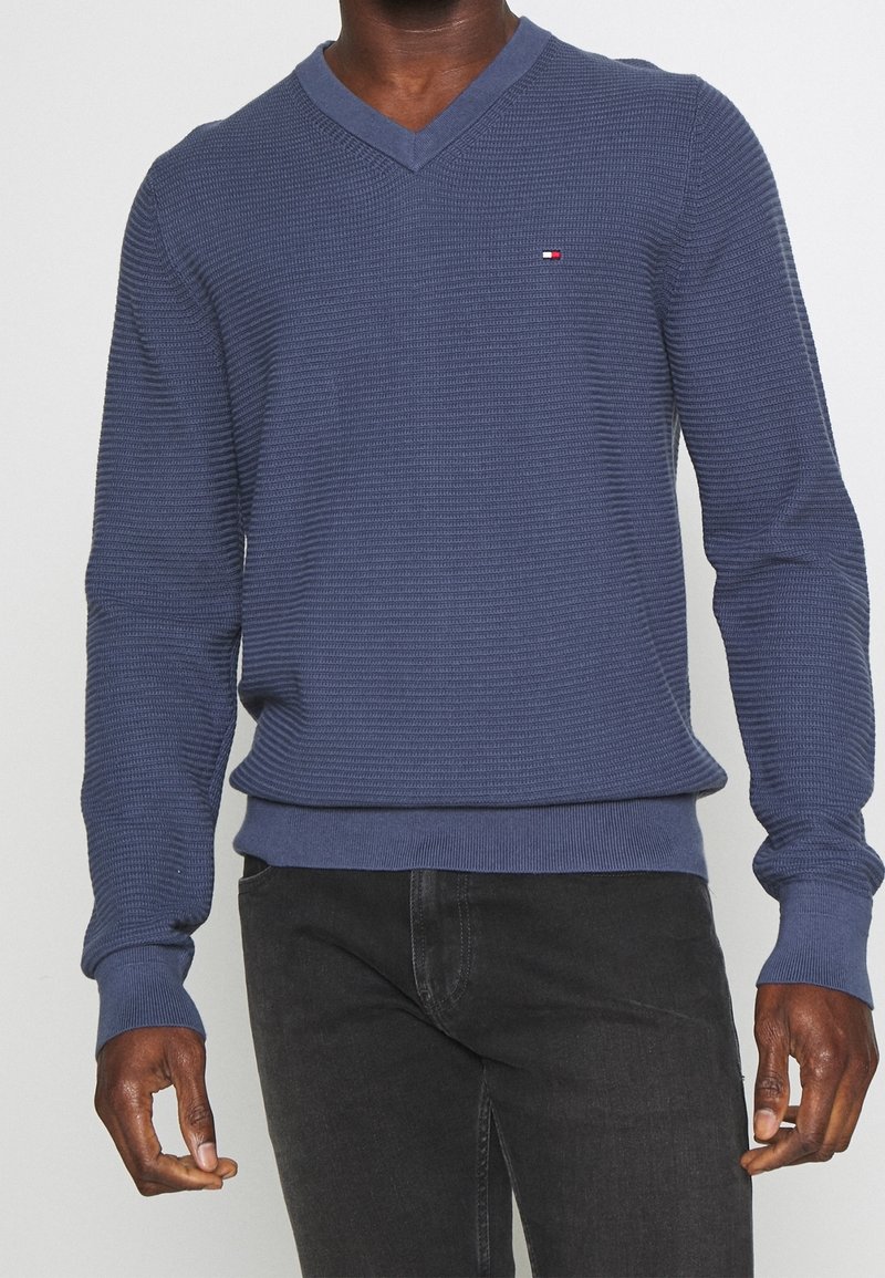 Homme portant un pull bleu texturé à col en V avec un petit logo sur la poitrine et un jean noir, debout devant un fond uni.