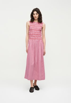 Kobieta z kręconymi włosami do ramion, ubrana w różowo-czerwoną sukienkę midi w paski bez rękawów oraz czarne, plecione buty wsuwane, stojąca na białym tle.