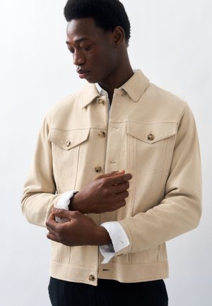 Jonge man die de mouwen van een beige jas met knopen en twee borstzakken rechtzet tegen een effen witte achtergrond