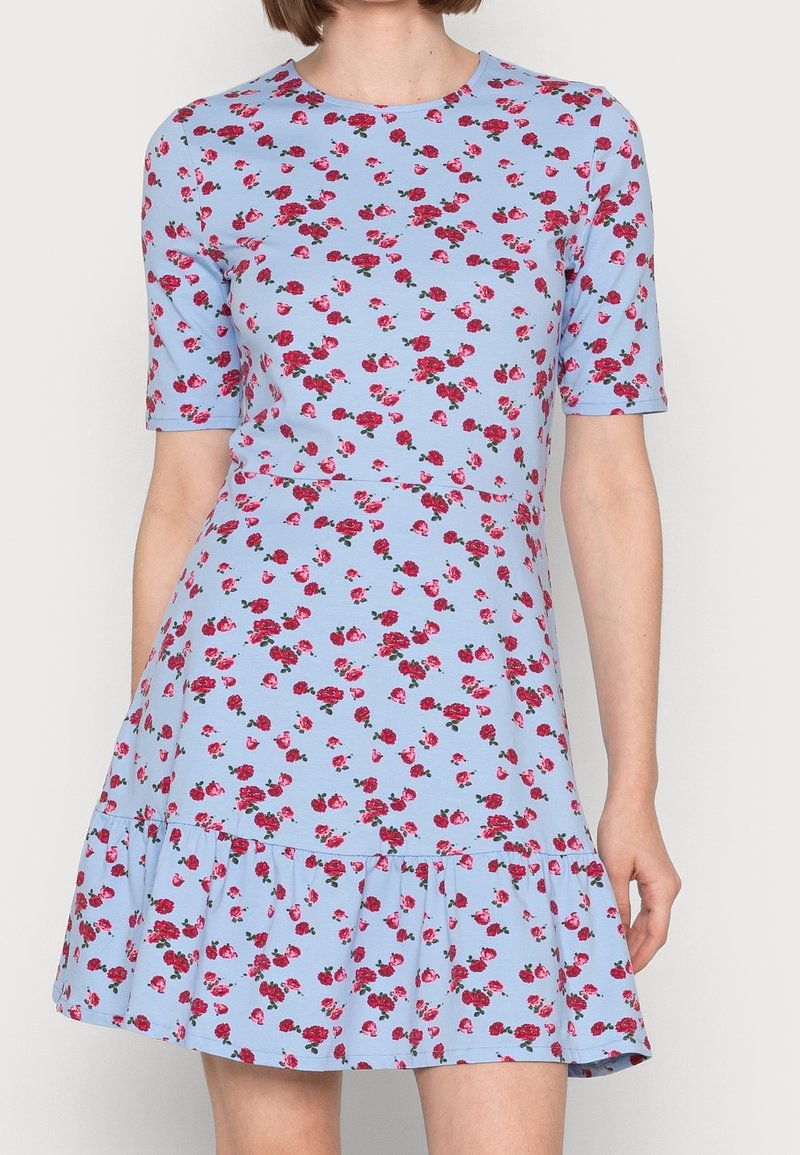 Robe bleu clair à manches courtes avec imprimé floral rouge, taille cintrée et ourlet à volants, portée par une personne debout.