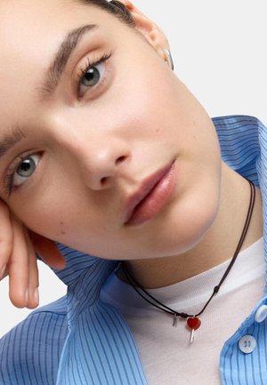 Close-up of a person with gray eyes, wearing a blue striped shirt, white top, silver hoop earring, and a black necklace with a red heart charm.