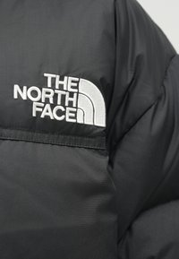 Crna prozračna jakna s bijelim izvezenim logotipom na ramenu, glatke teksture i slojevitim dizajnom s mat završetkom.