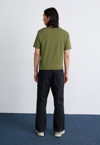 Een persoon met lang haar, in een olijfgroene shirt en zwarte broek, staat met de rug naar een effen muur op een blauwe tapijt.