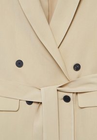 Gros plan sur un manteau beige croisé, boutons noirs et ceinture assortie en tissu nouée à la taille.