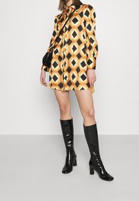 Robe chemise avec un motif géométrique noir et or ; manches longues et fentes sur les côtés. Le modèle porte des bottes noires en vernis arrivant aux genoux.