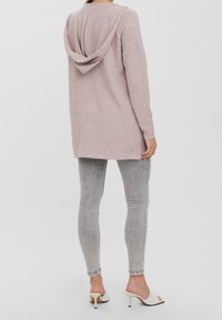 Lichtroze capuchonvest gemaakt van zachte, gestructureerde stof, met lange mouwen en een langere achterkant, gecombineerd met grijze skinny jeans.