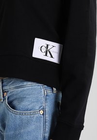 Czarna bluzka z długim rękawem z białą naszywką z logo Calvin Klein Jeans, zestawiona z jasnoniebieskimi dżinsami, ukazująca talię i biodra.