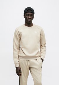 Béžová mikina s okrúhlym výstrihom a rebrovanými manžetami, na hrudi malý biely logo Adidas. Štýlovo doplnená o béžové nohavice v rovnakom odtieni.