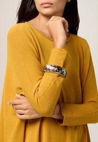 Un pull en tricot jaune avec des poignets côtelés, arborant un grand bracelet jonc marbré noir et crème brillant au poignet du modèle.
