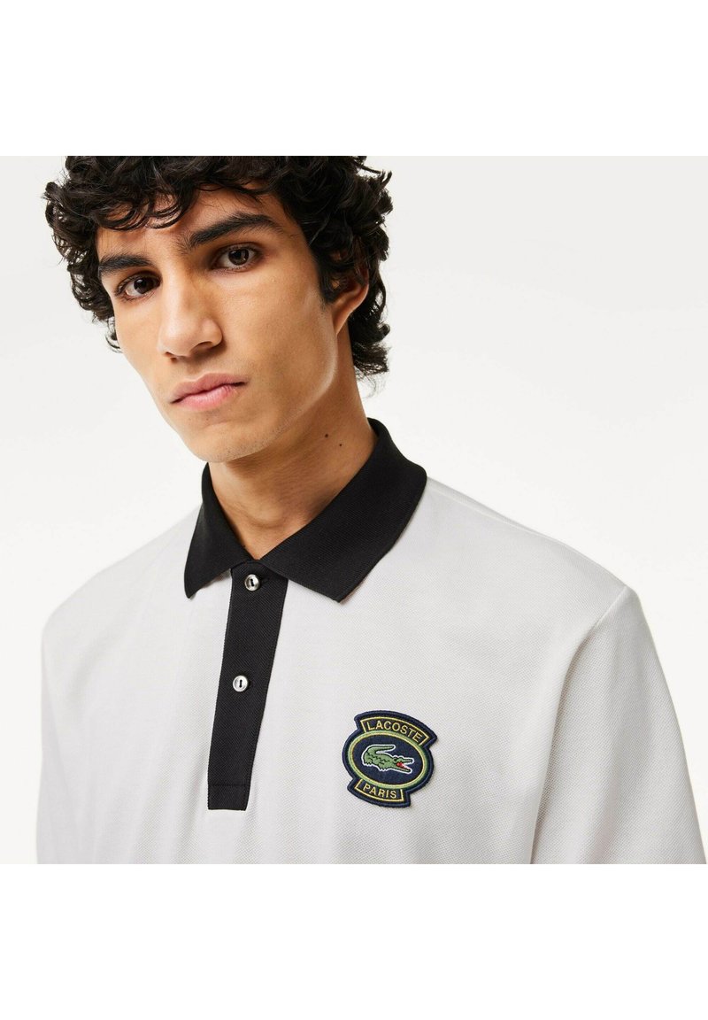 Polo-shirt blanc avec un col noir, arborant un logo Lacoste brodé vert et bleu sur la poitrine. Le matériau semble texturé et respirant.