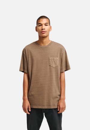 Junger Mann, der ein lockeres braunes T-Shirt mit Brusttasche und schwarze Hosen vor einem schlichten hellen Hintergrund trägt.