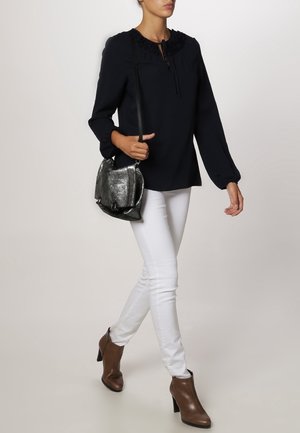 Blusa blu navy con scollatura arricciata e dettaglio con fiocco, abbinata a jeans skinny bianchi e stivaletti marroni. La modella porta una borsa metallica.