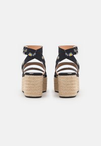 Een paar zwarte platformsandalen met stofbanden met madeliefjesprint en gewoven touwzolen, gezien van achteren op een witte achtergrond.