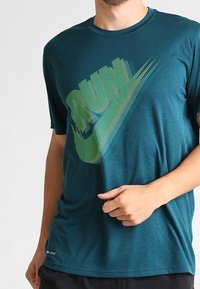 Teal atletisk T-shirt med en stor grön och marinblårandig Nike-logga på framsidan, tillverkad av lätt, andningsbart material.
