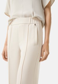 Lichtbeige op maat gemaakte broek van een gladde stof, met een relaxte tailleband, zijzakken en subtiele plooidetails.