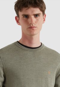 Zielona oliwkowa dzianinowa sweter z okrągłym dekoltem i prążkowanymi brzegami. Posiada małe pomarańczowe logo na lewej piersi. Miękka tekstura i lekka.