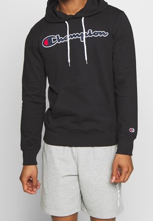 Hoodie noir en coton avec des cordons blancs, poche avant et logo Champion brodé en bleu et rouge. Associé à un short gris.