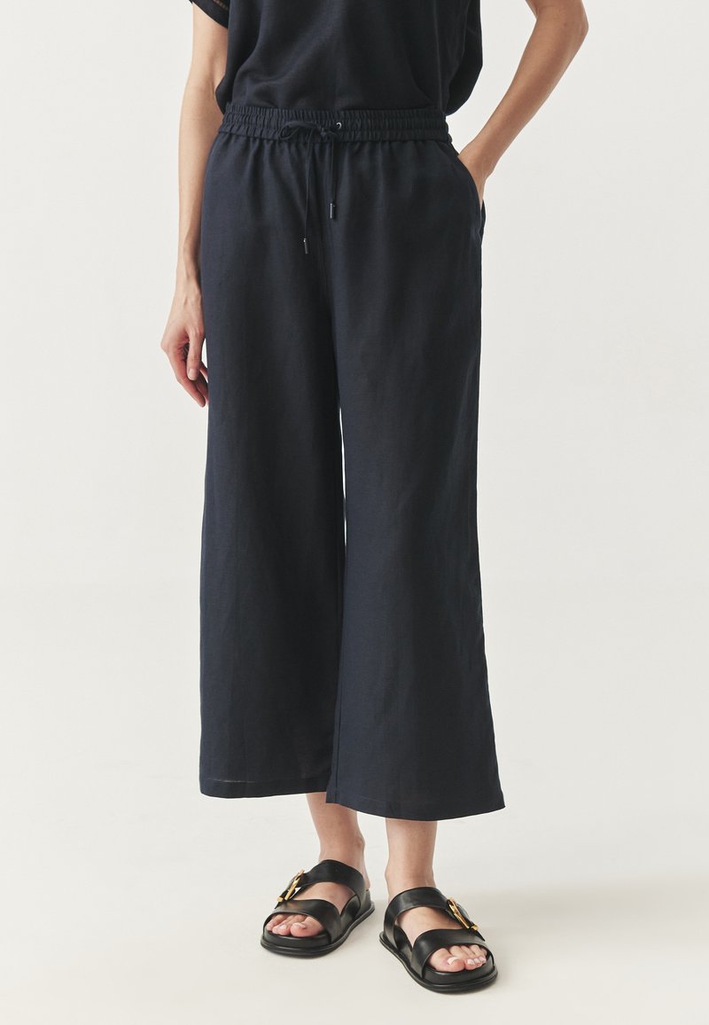 Marineblauwe wijde broek van lichtgewicht stof, met een elastische tailleband met trekkoord en zijzakken. Gecombineerd met zwarte platte sandalen.