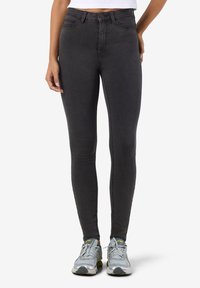 Juodos spalvos siauri džinsai su aukštu juosmeniu, sklandžios džinsinės tekstūros ir minimaliais siuvimo detalėmis, derinami su pilkais sportiniais batais.