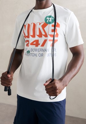 Osoba držící černé švihadlo, oblečená v bílém tričku Nike 24/7 s grafikou a tmavě modrých šortkách.