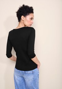 Musta, tiukasti istuva pitkähihainen paita, joka on valmistettu joustavasta materiaalista, yhdistettynä vaaleansinisiin, löysästi istuviin farkkuihin; aihe kääntyy pois kamerasta.