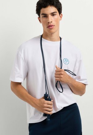 Junger Mann in weißem T-Shirt hält ein schwarzes Springseil, das um seinen Hals gelegt ist, vor einem schlichten hellen Hintergrund.